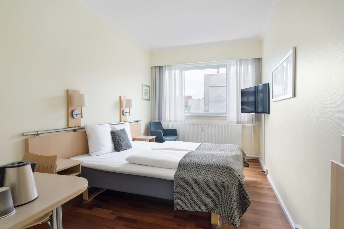 Une chambre d’hôtel comprenant un lit, une table de chevet, une lampe, une chaise, une télévision et une fenêtre avec des rideaux.