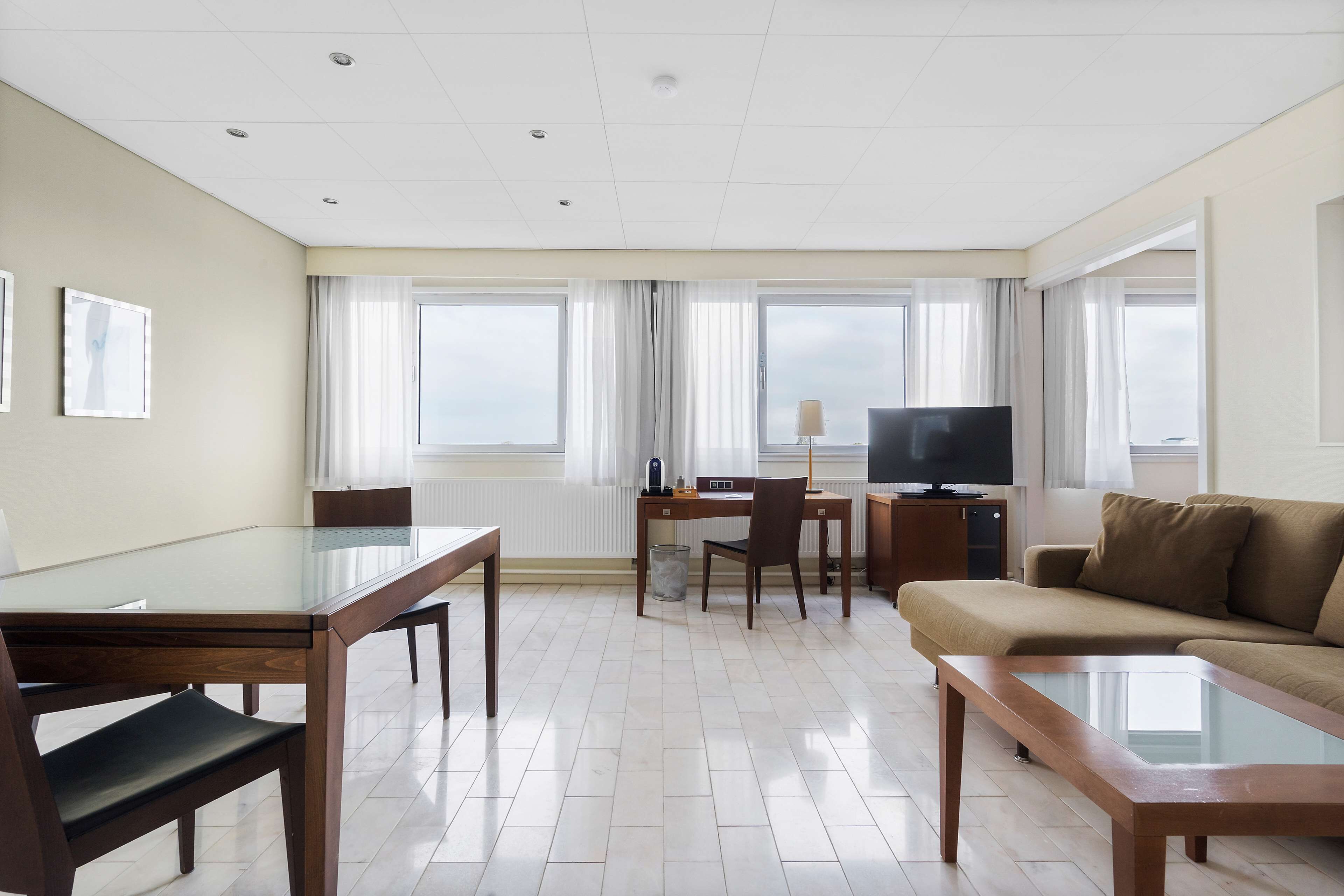 A modern hotel room with a dining table, a sofa, a TV, and large windows with curtains.