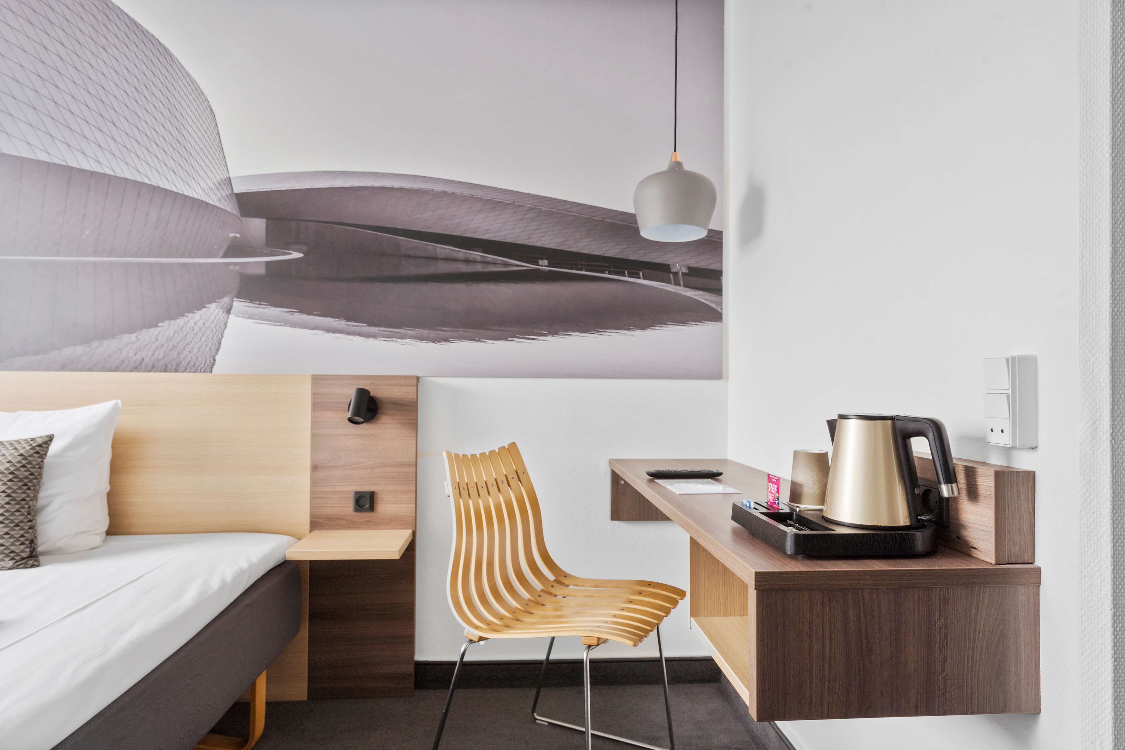Habitación de hotel moderna con una cama, una silla de madera, un escritorio con una tetera y un diseño de techo único.