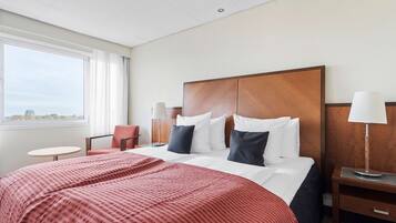 A hotel room with a large bed, a red bedspread, a wooden headboard, a nightstand with a lamp, a chair, and a window with a view of the sky.