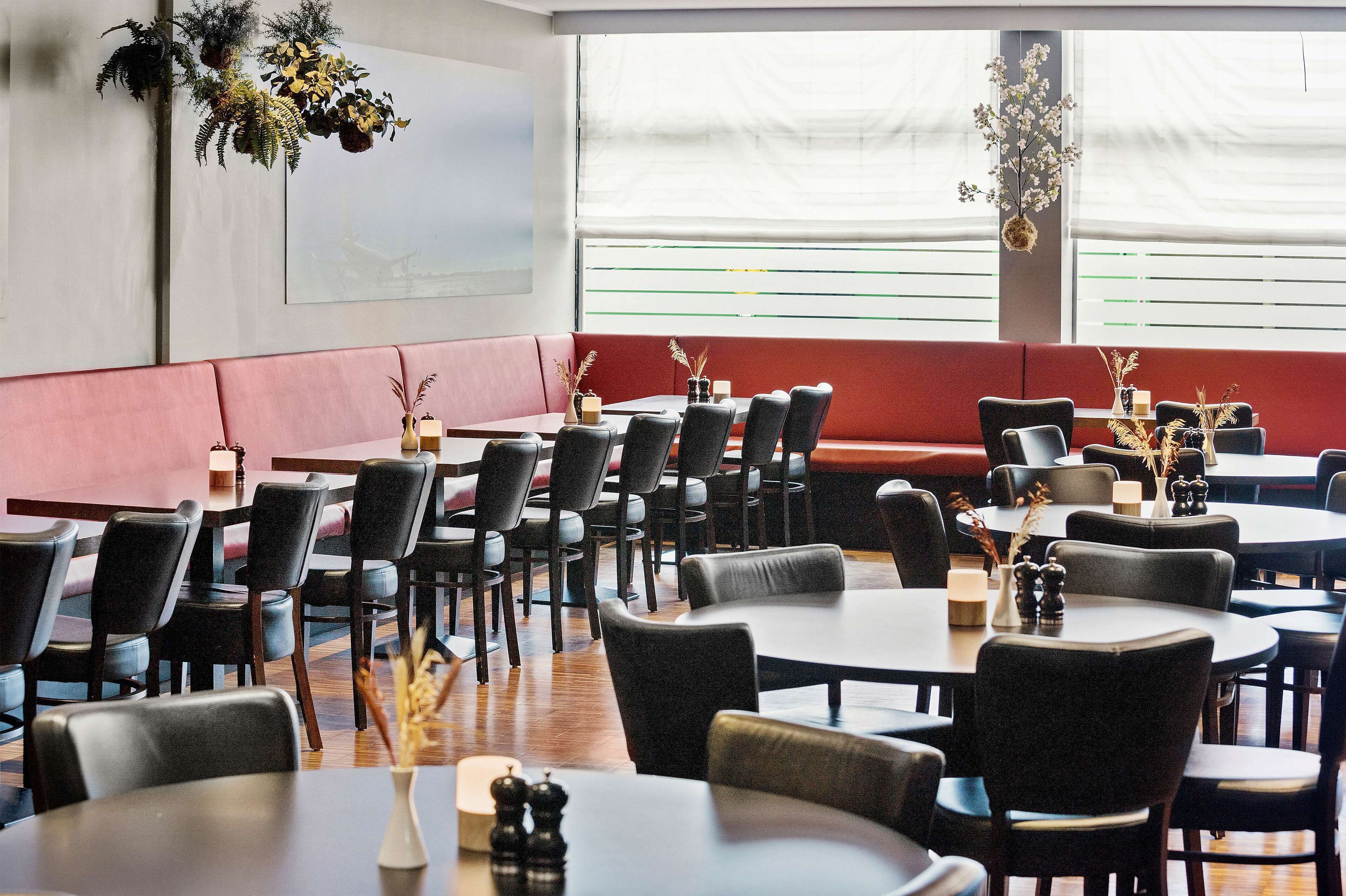 Een restaurant met meerdere tafels en stoelen, een rode, beklede bank en decoratieve planten.