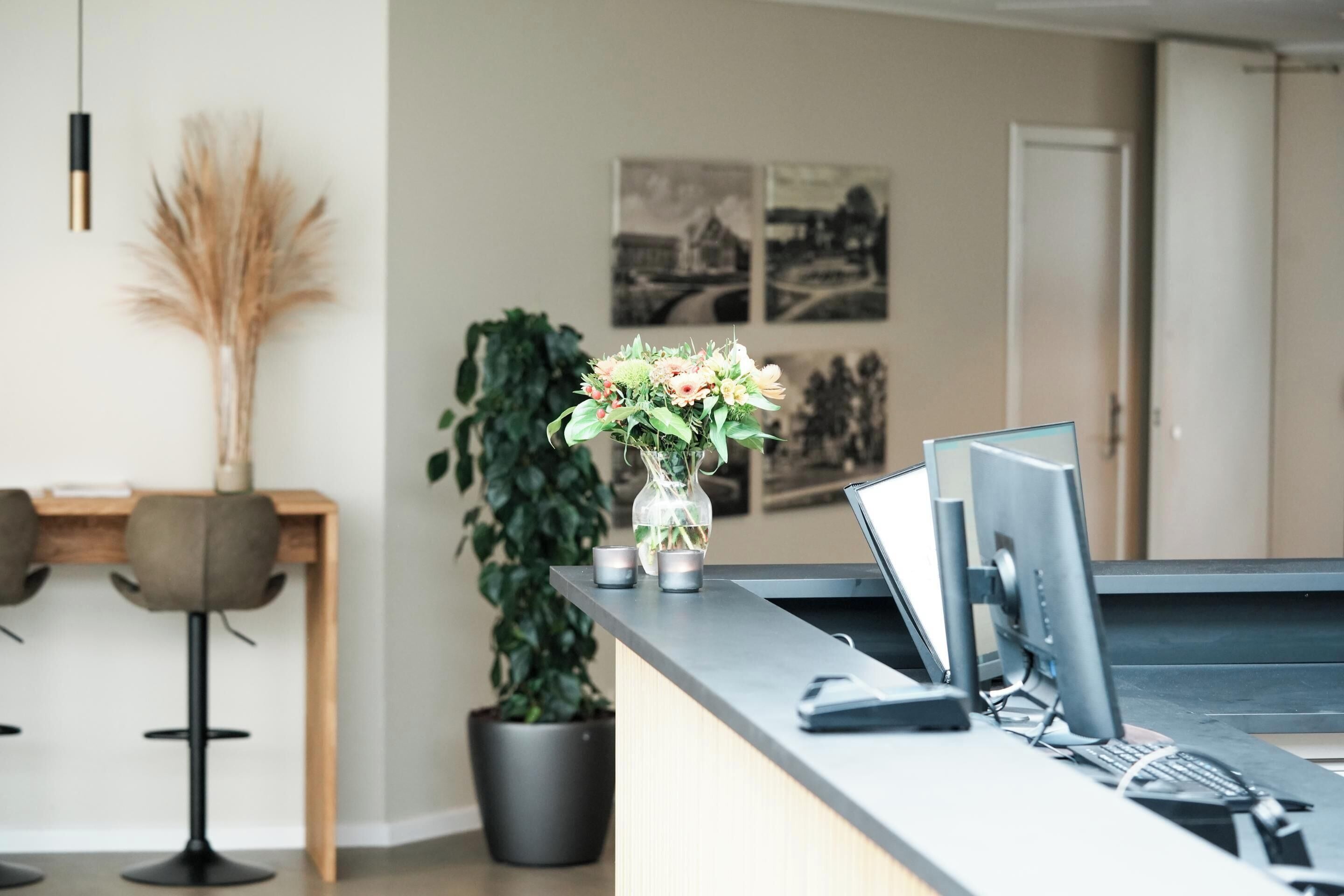En reception med en bærbar computer, en skærm og en vase med blomster. To stole og en potteplante ses i baggrunden.