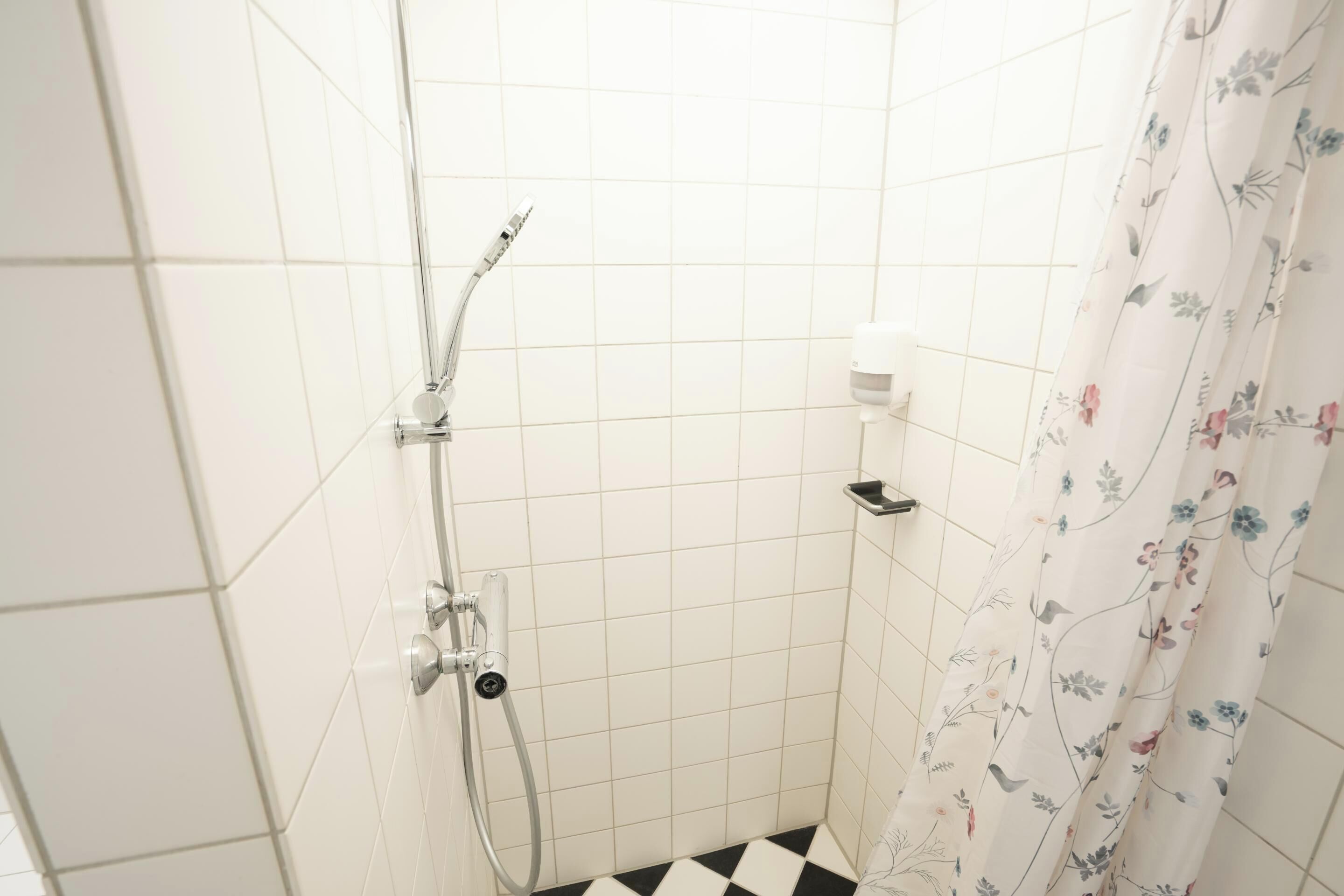 Un petit espace de douche carrelé, équipé d’un pommeau de douche et d’un rideau de douche à motifs floraux.