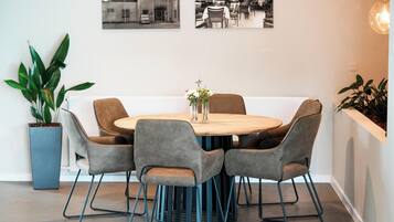 A dining area with a round wooden table, six grey upholstered chairs, and a vase of flowers. There are two black-and-white photographs on the wall above the table.