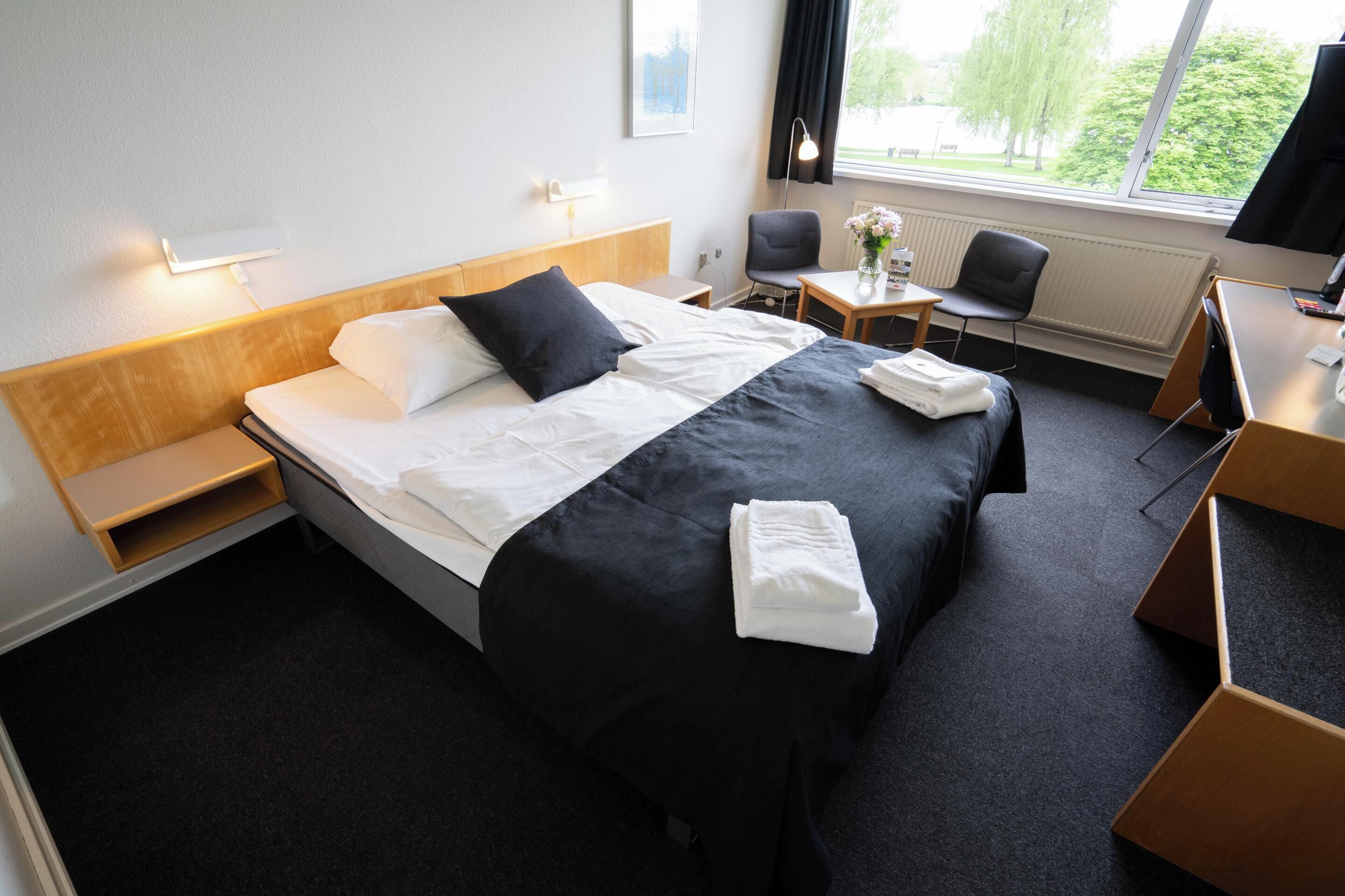 A hotel room with a bed, a desk, a chair, and a window with a view of trees.