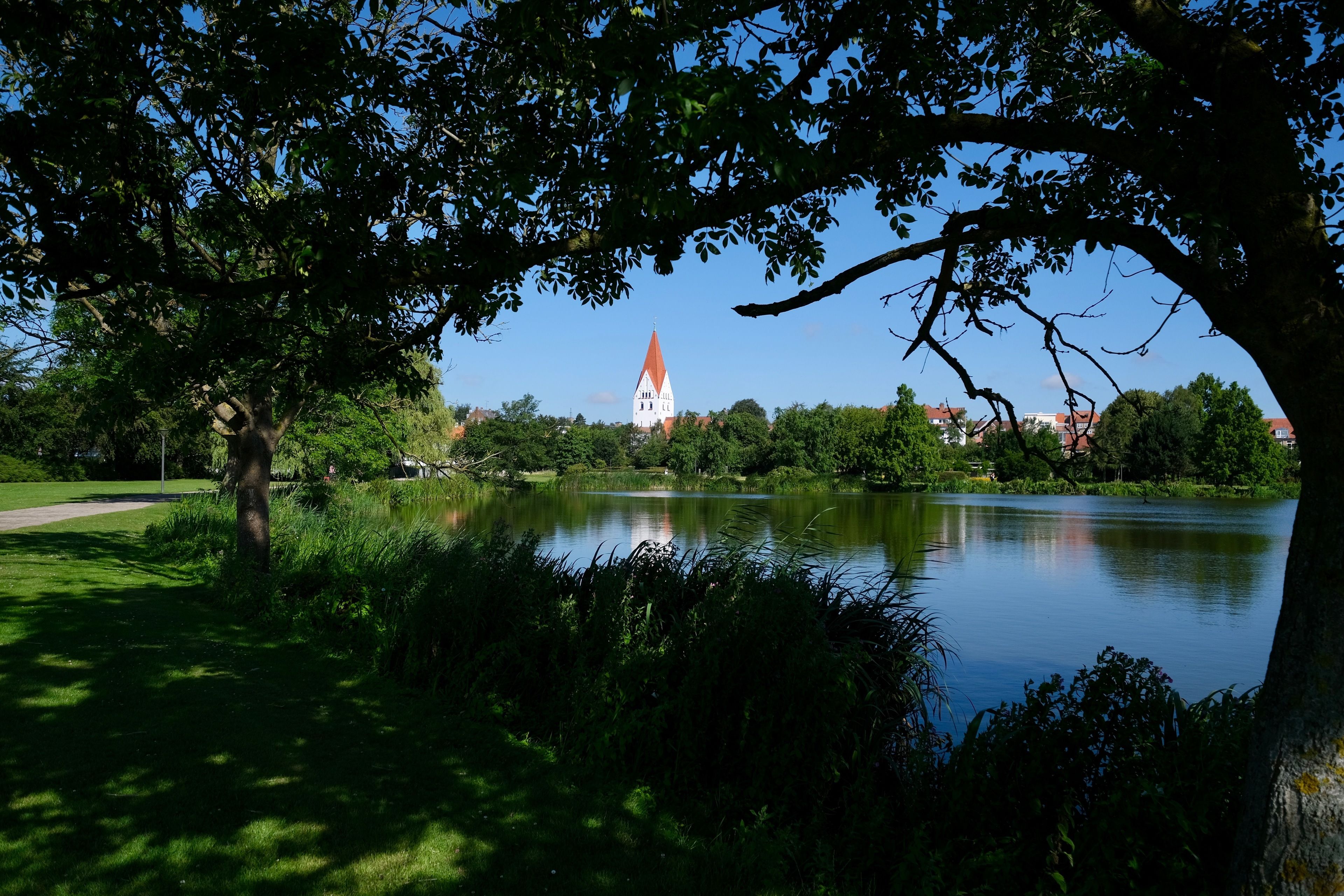 A serene park with a lake, lush greenery, and a distant church tower.