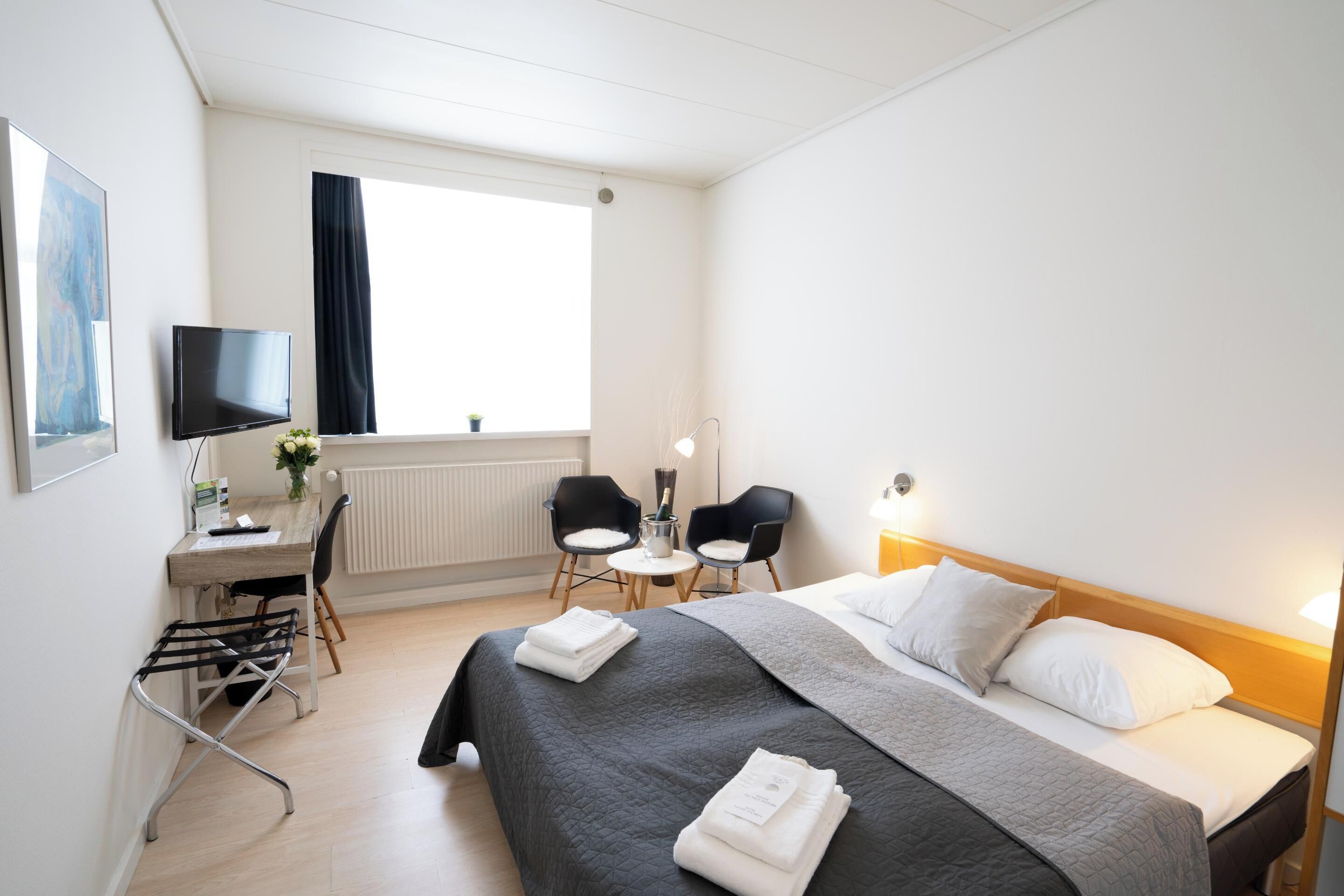 Hotellihuone, jossa on sänky, työpöytä, kaksi tuolia, televisio ja ikkuna.