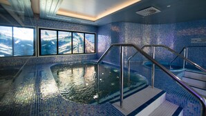 Una piscina interior con suelo y paredes de azulejos, un ventanal de cristal y una escalera de metal.