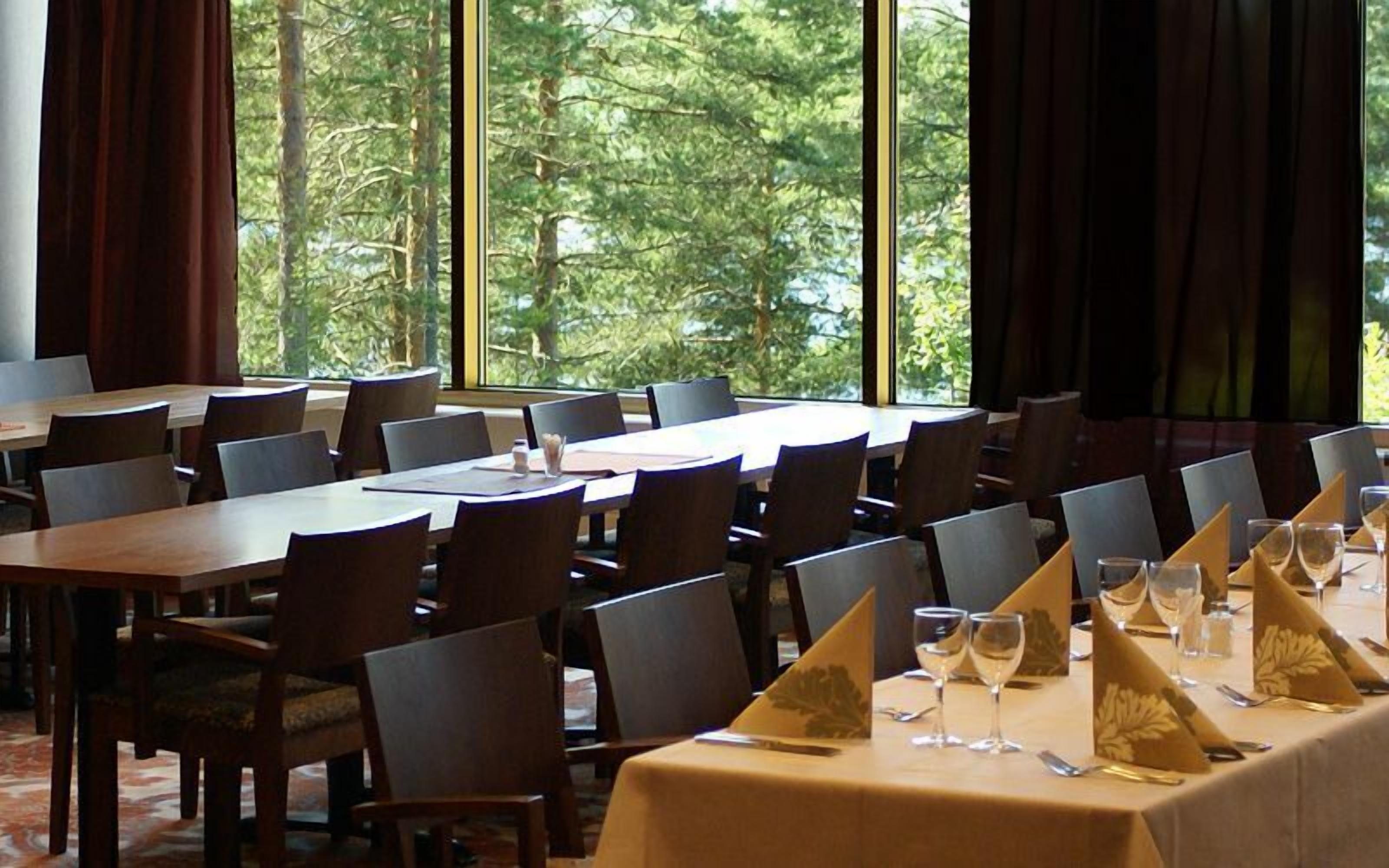 A conference room with multiple tables and chairs, large windows with a view of trees, and a carpeted floor.