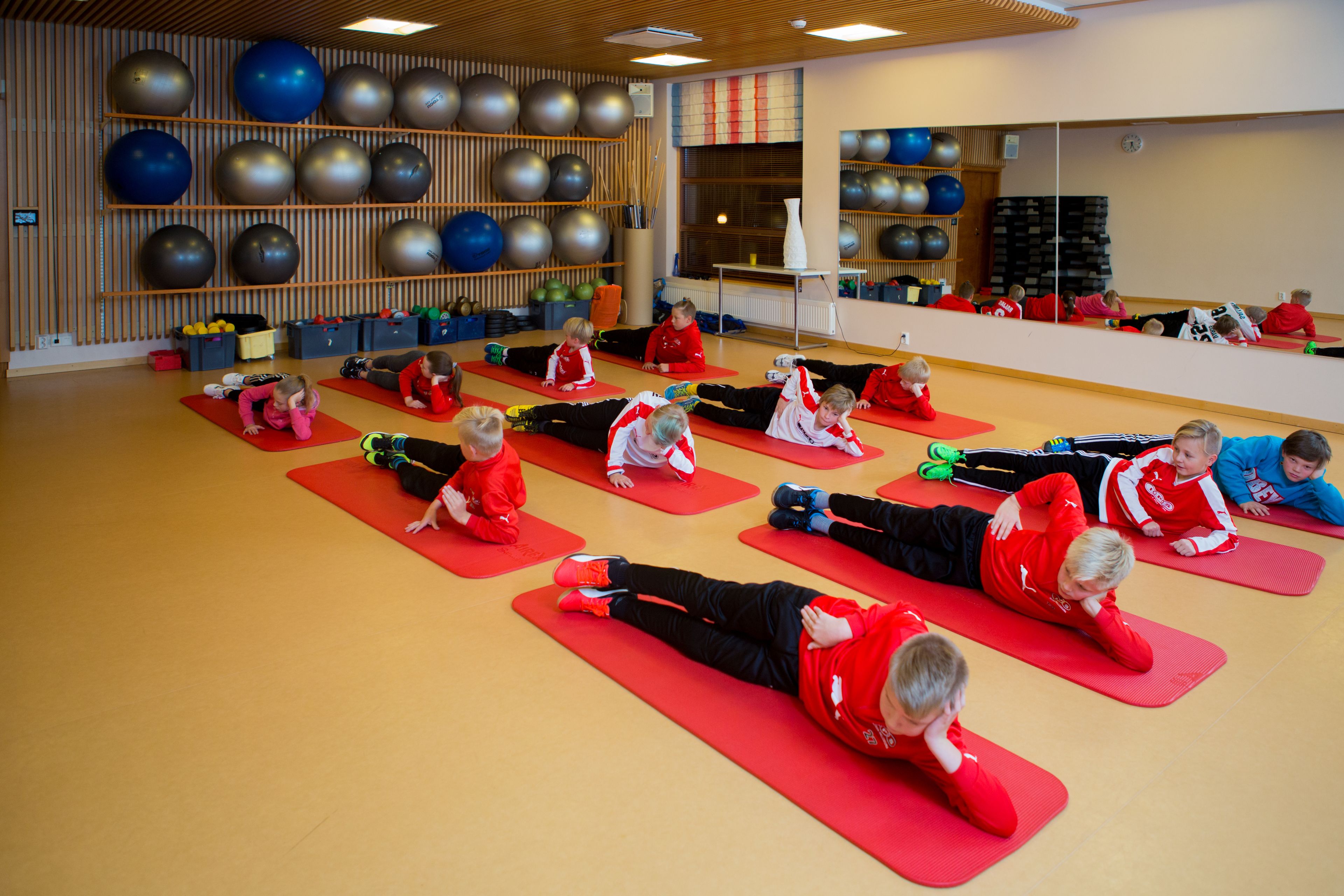 A group of people in red shirts doing a stretching exercise on red mats in a room with exercise balls on the walls.