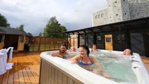 Un jacuzzi entouré de personnes, une terrasse en bois et un bâtiment en pierre avec un moulin à vent en arrière-plan.