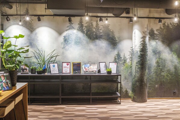 A reception area with a wooden desk, a plant, framed certificates, and a wall mural of trees. - Original Sokos Hotel Koljonvirta (Iisalmi)