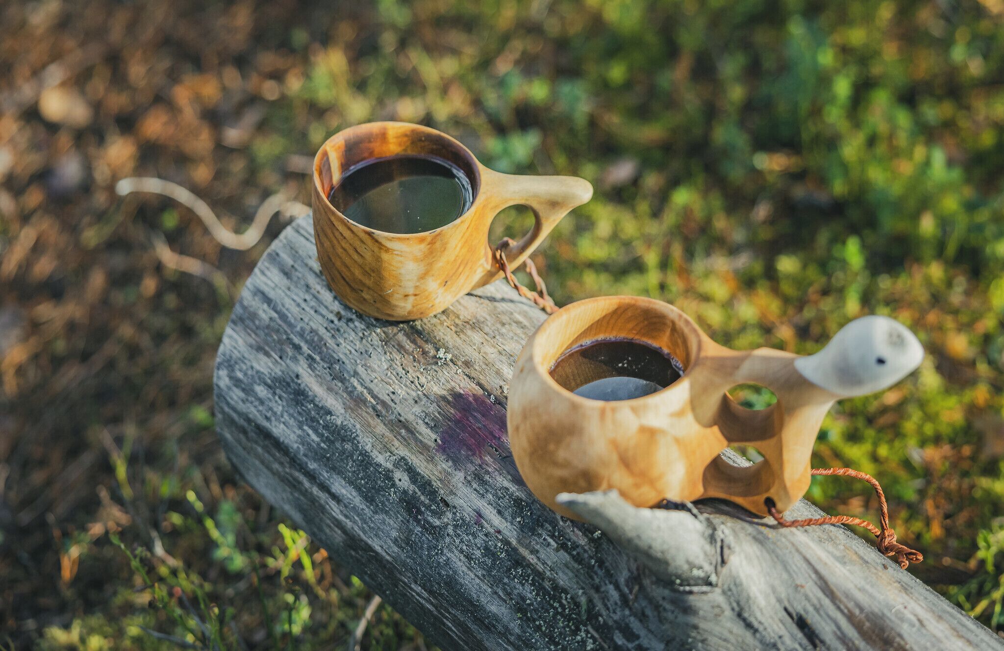 Two wooden cups with handles, filled with a dark liquid, placed on a wooden log.