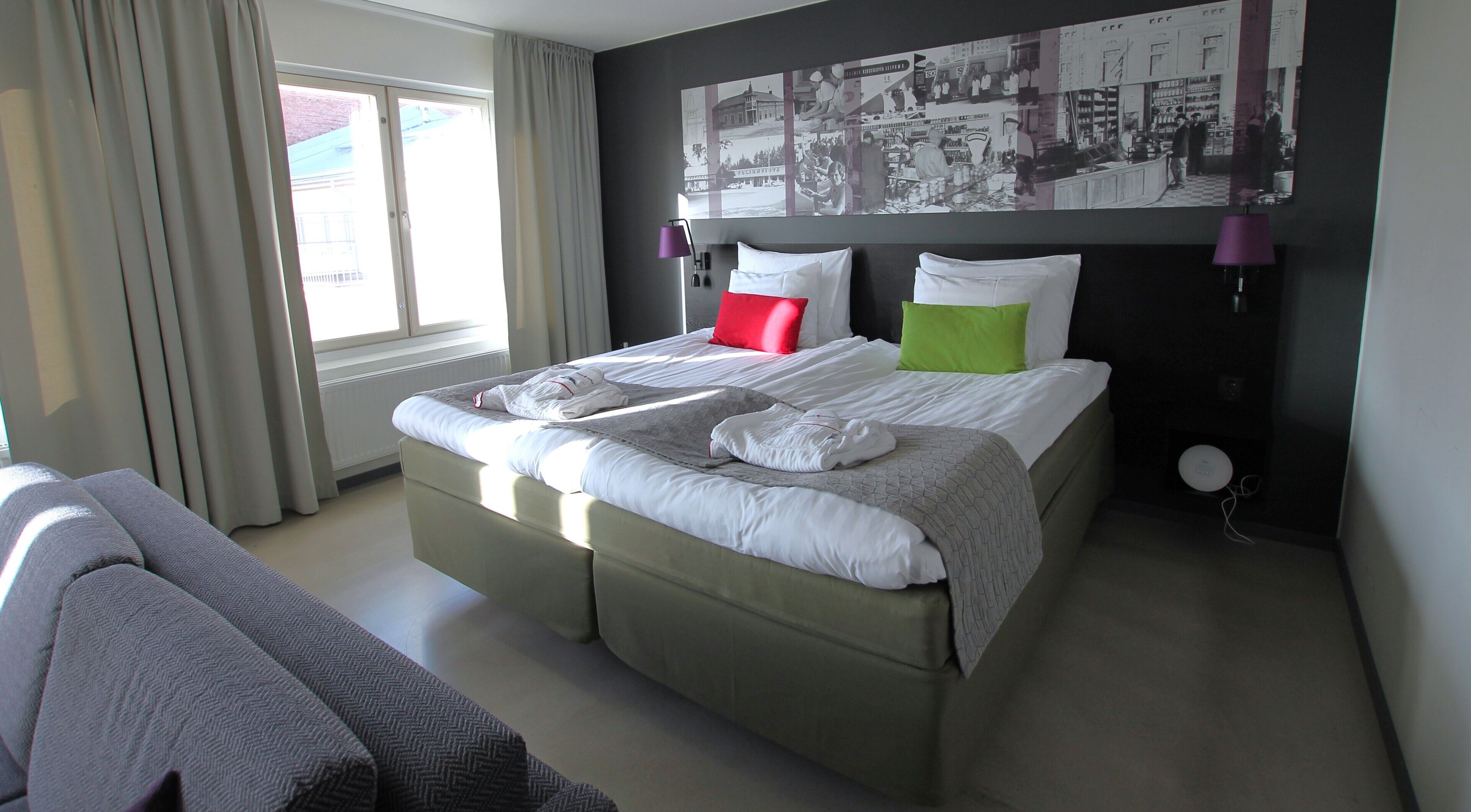 A neatly made bed with pillows, a bedside table with a lamp, and a large window with curtains.