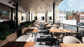 Une terrasse extérieure avec des tables en bois et des chaises en osier, entourée de verdure et surplombée d’un plafond en bois agrémenté de guirlandes lumineuses.