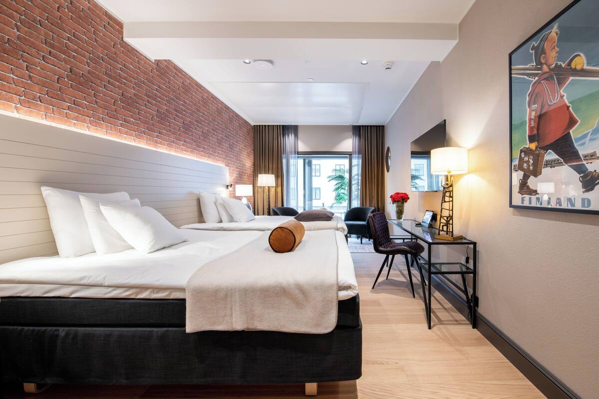 Habitación de hotel con pared de ladrillo, dos camas, un escritorio con silla y un cuadro en la pared.