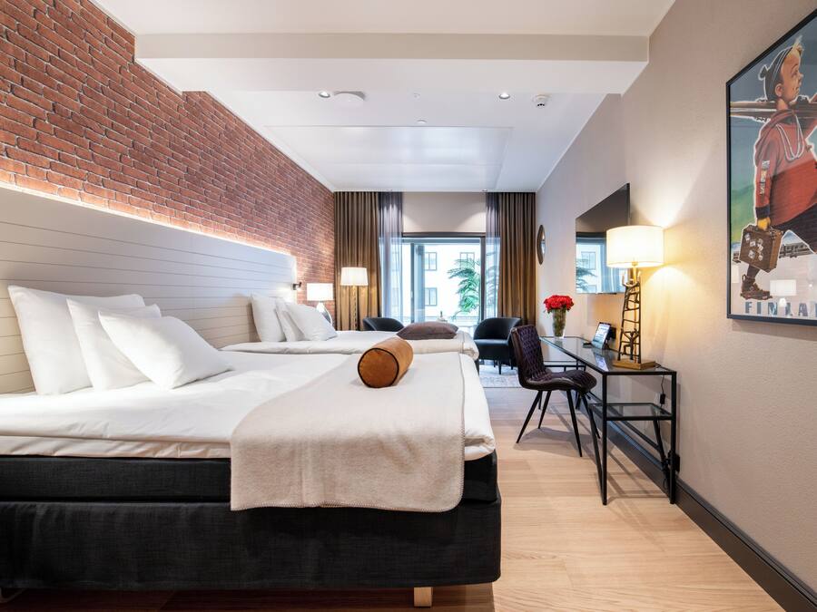 A hotel room with a brick wall, two beds, a desk with a chair, and a painting on the wall.