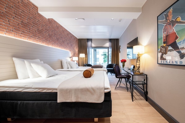 A hotel room with a brick wall, two beds, a desk with a chair, and a painting on the wall.