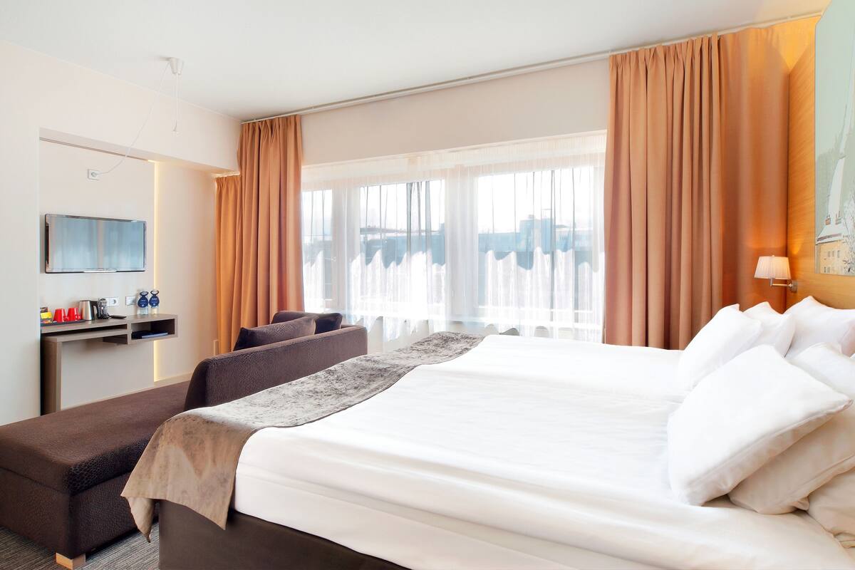 Una habitación de hotel con una cama grande, un escritorio y un televisor de pantalla plana, y una ventana con cortinas.