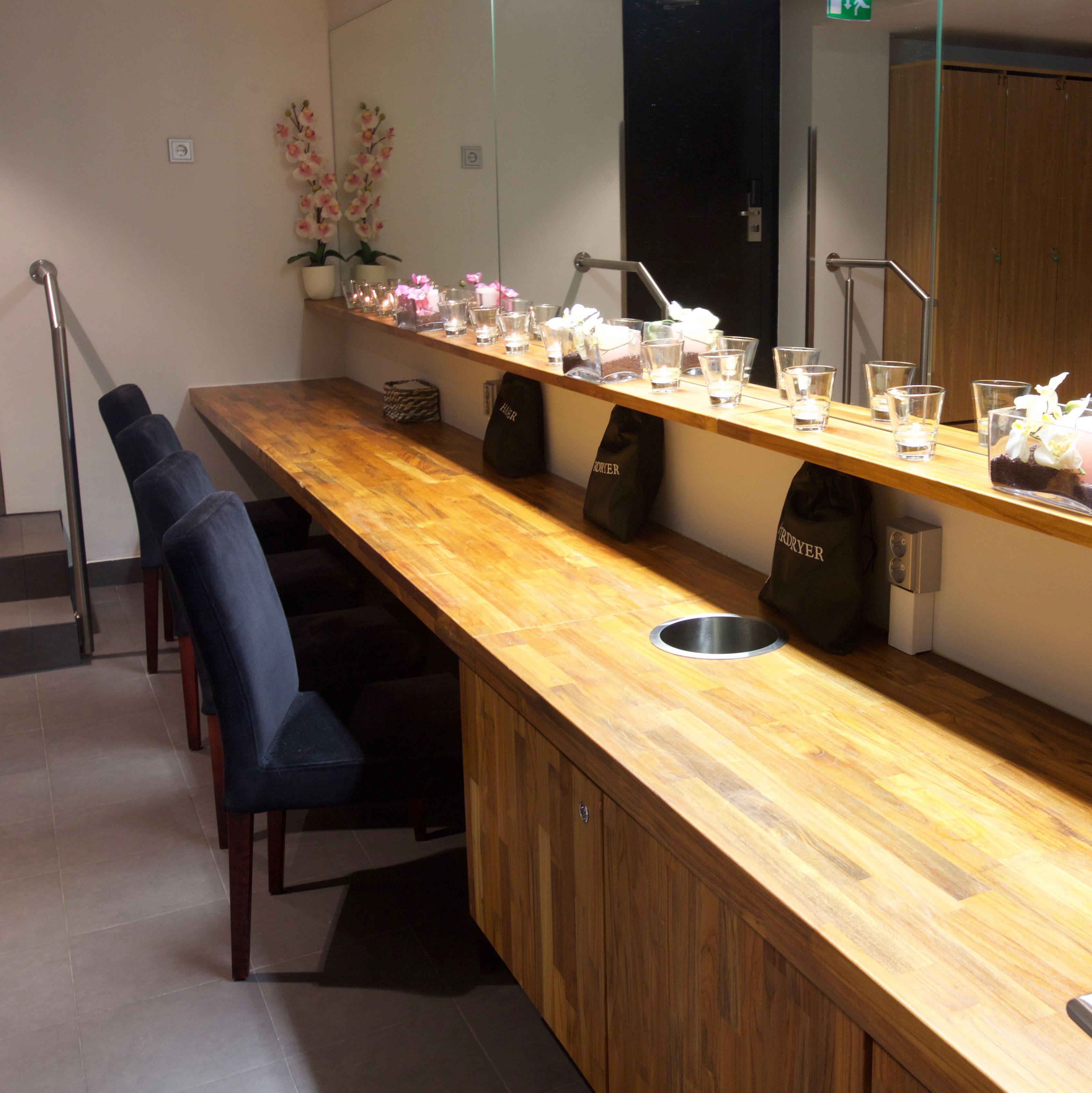 A long wooden counter with multiple glass tumblers, a metal sink, and a mirror above. There are chairs along the wall and a staircase to the left.