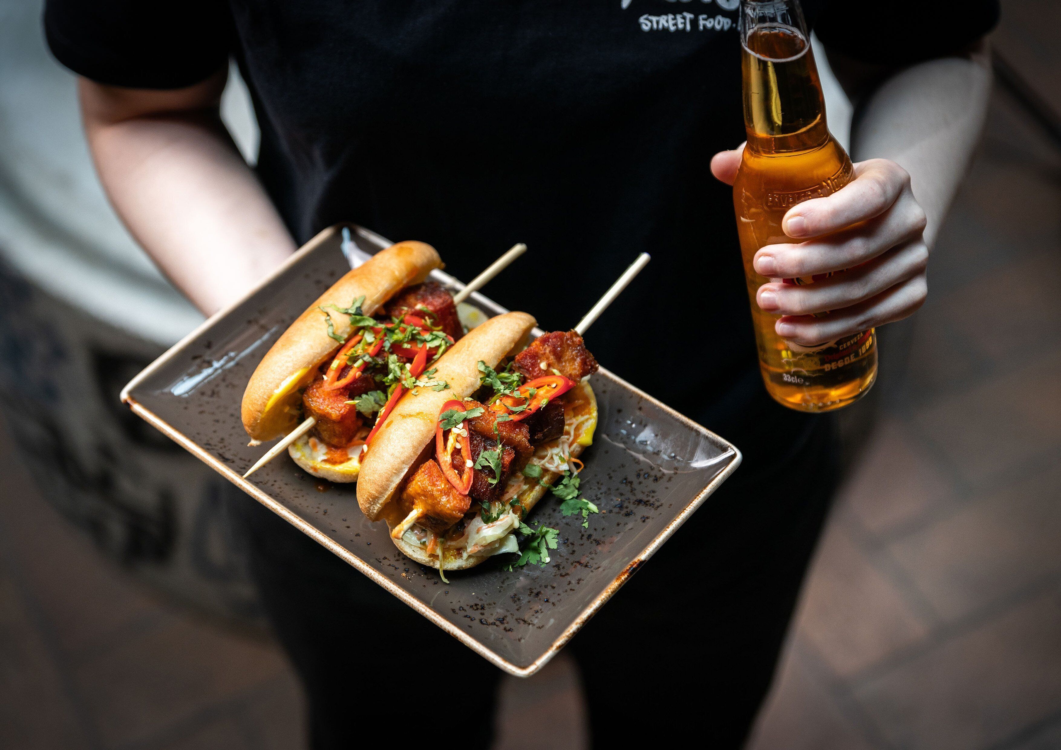 A person holding a tray with two skewers of food and a bottle of beer.