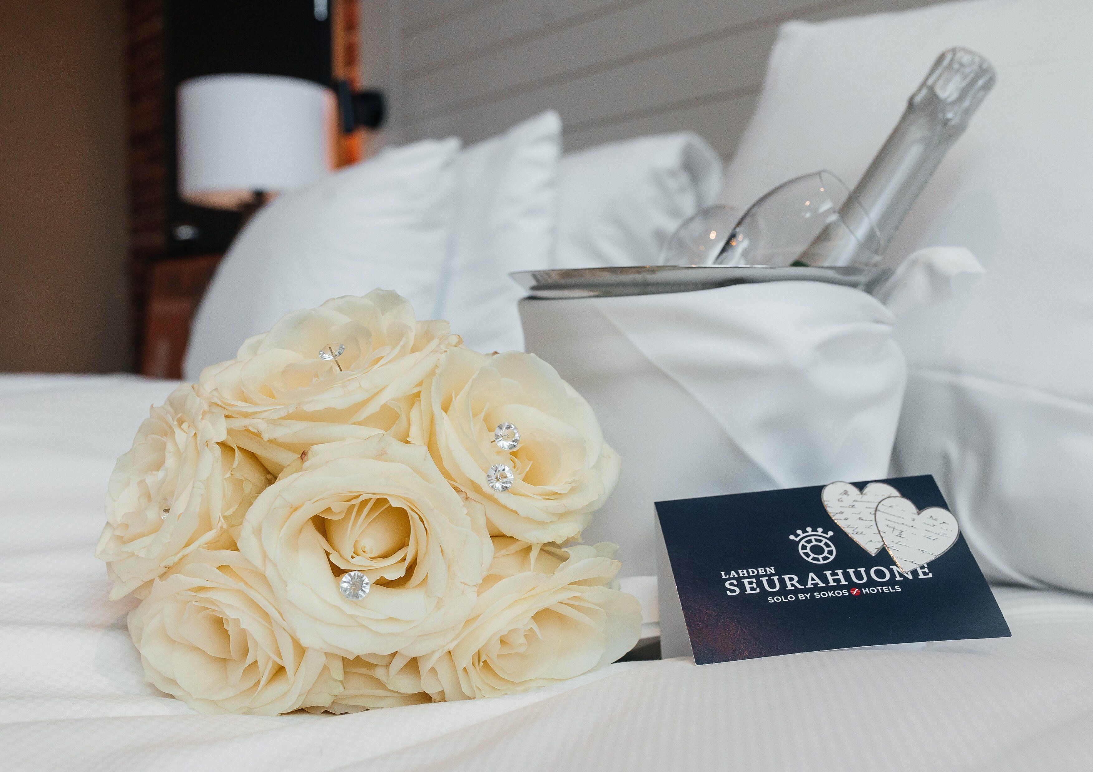 A bouquet of white roses with a diamond, a glass of champagne, and a business card on a bed.