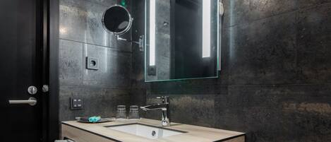 Un baño moderno con pared de baldosas oscuras, tocador de madera, espejo grande y lámpara colgante.
