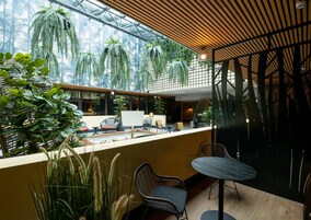 A modern indoor space with a glass ceiling, allowing natural light to illuminate the area. There are several chairs and a table, suggesting a seating area.