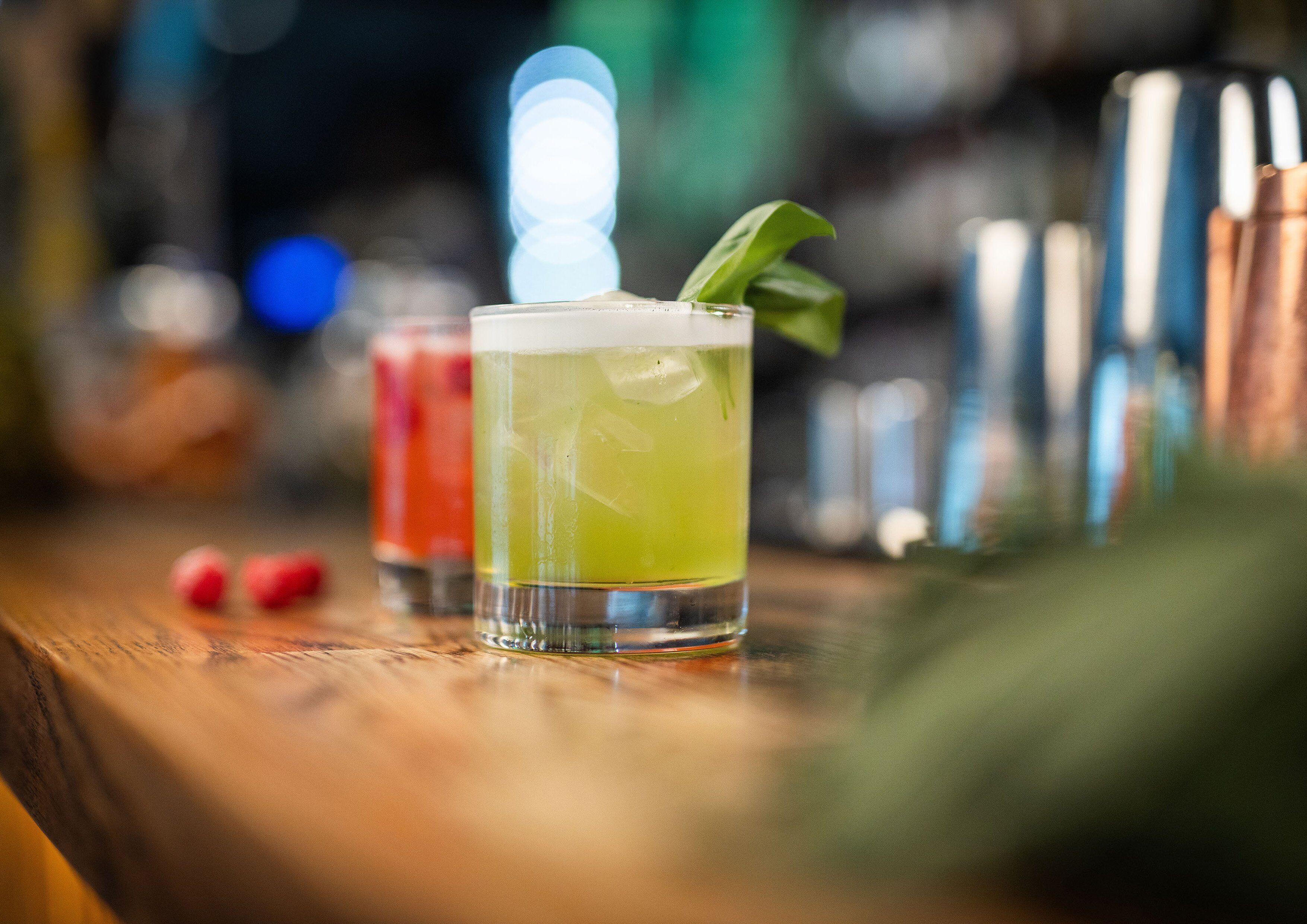 Baaritiski, jossa on lasi vihreää cocktailia, jonka päällä on koristeena lehti. Taustalla on epätarkka kuva baaritarvikkeista, ja etualalla on puinen pinta.