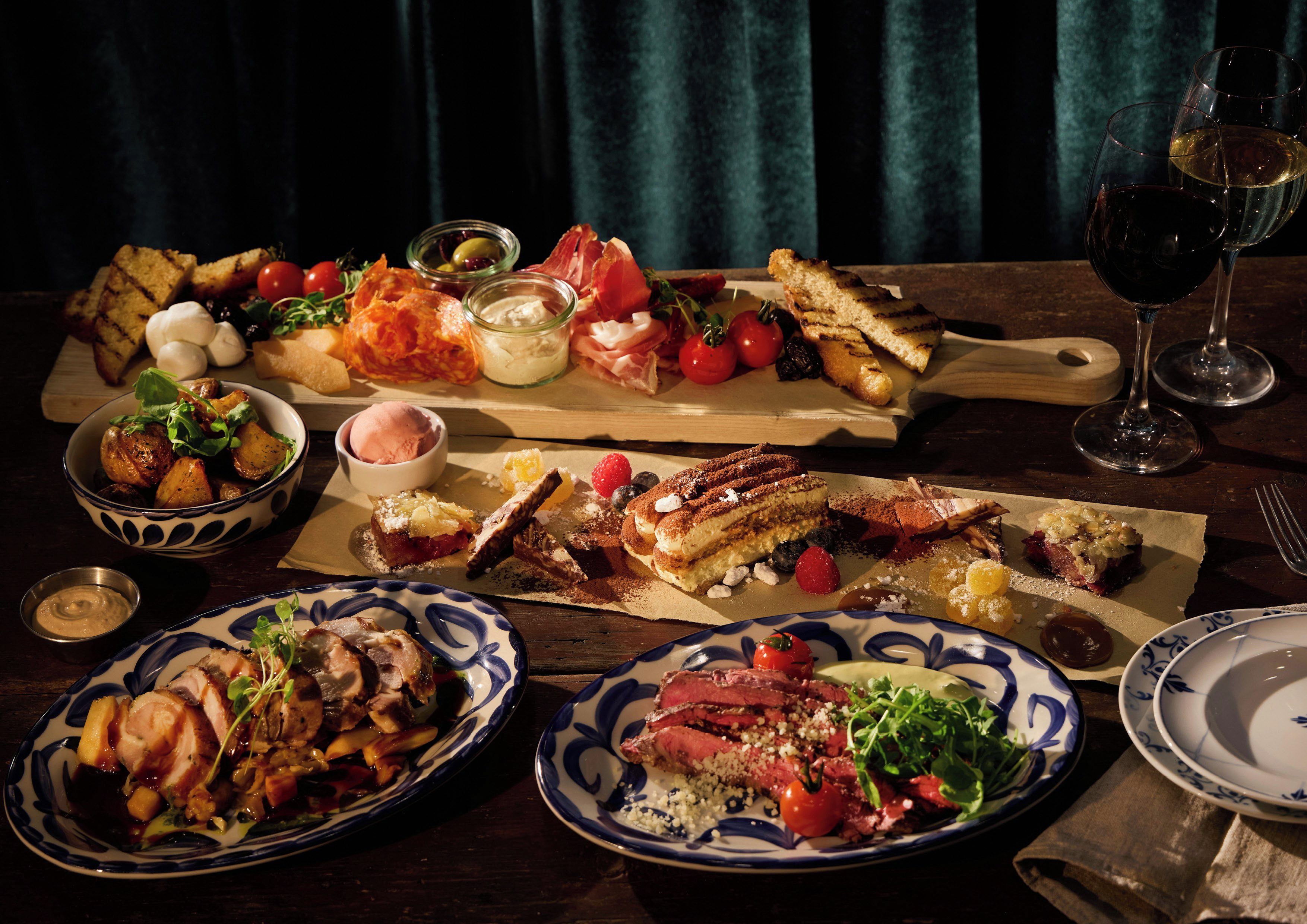 A variety of dishes on a wooden table, including meats, vegetables, and desserts, with wine glasses and a small bowl of sauce.
