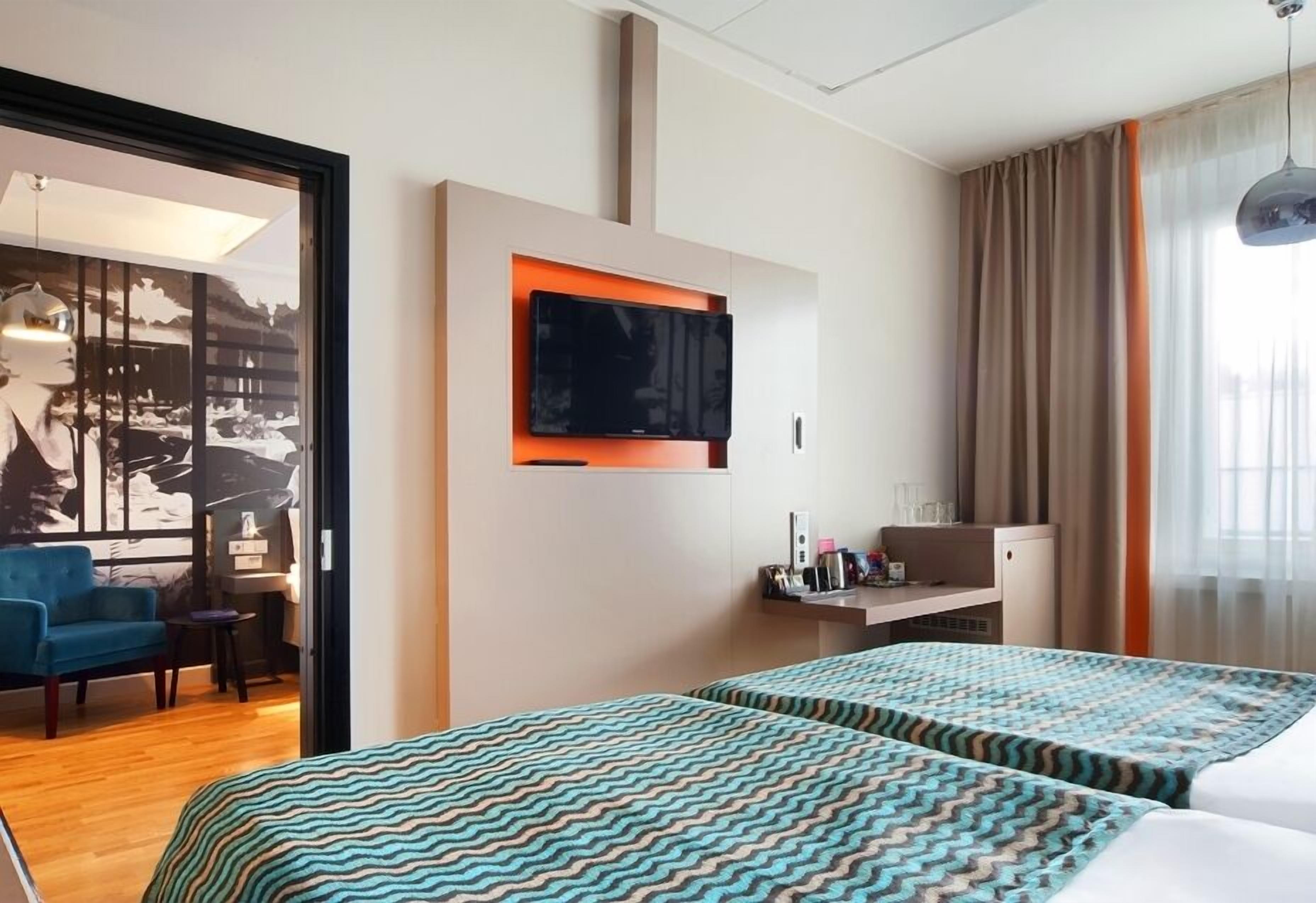 Ein Hotelzimmer mit zwei Betten, einem an der Wand montierten Fernseher, einem Schreibtisch mit Stuhl und einem Vorhangfenster.