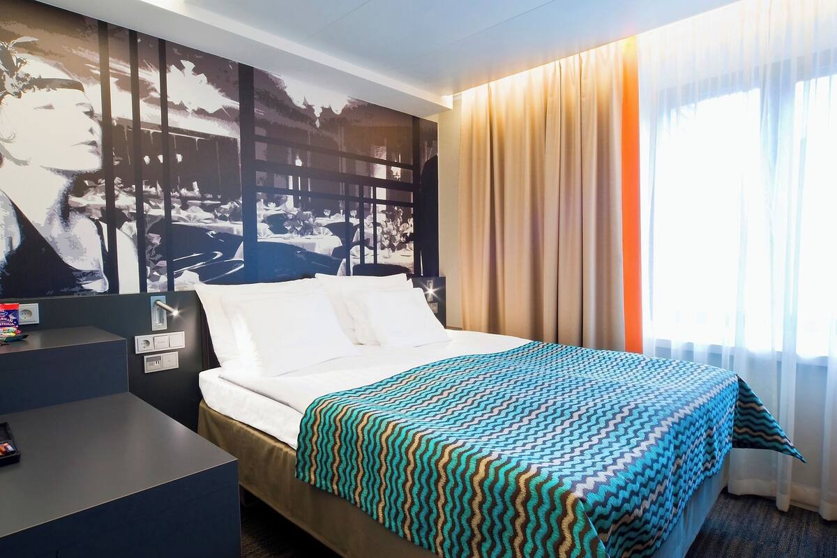 Habitación de hotel con cama, mesita de noche, televisor montado en la pared y ventana con cortinas.