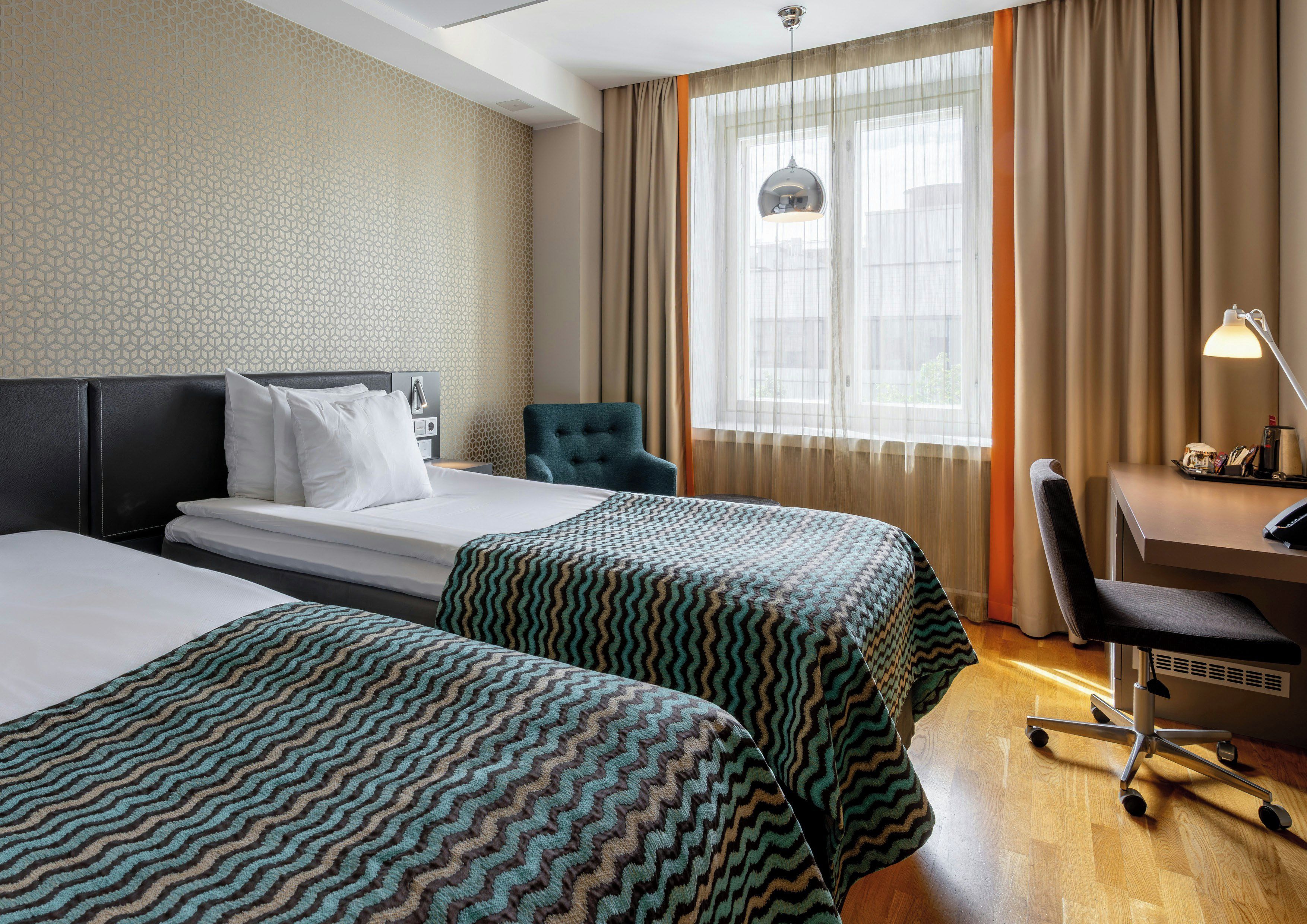 Ein Hotelzimmer mit zwei Betten, einem Schreibtisch, einem Stuhl und einem fensterbepflanzten Fenster.