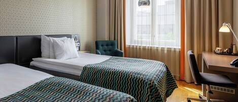 Ein Hotelzimmer mit zwei Betten, einem Schreibtisch, einem Stuhl und einem fensterbepflanzten Fenster.