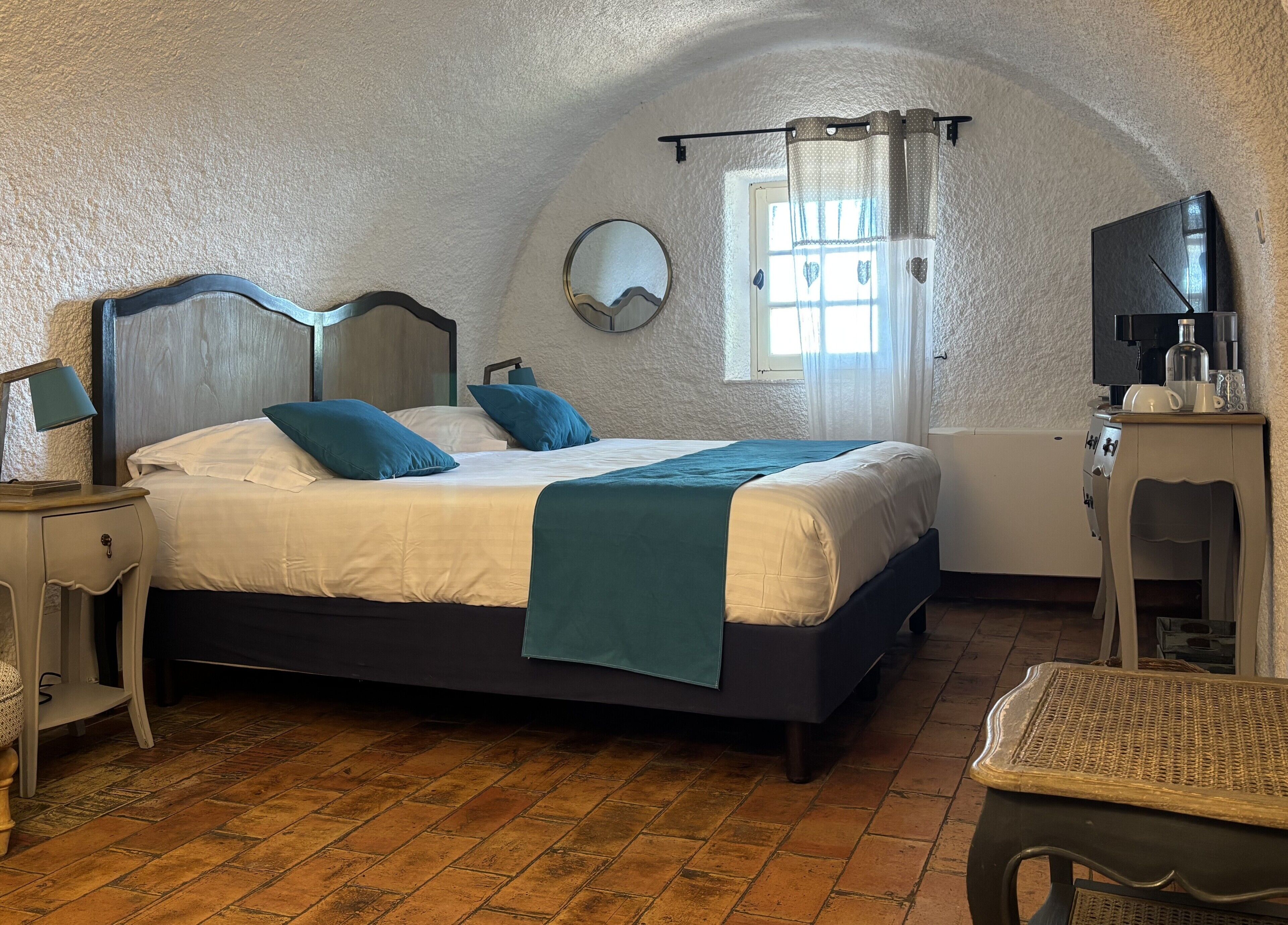 Un dormitorio con cama, mesitas de noche, espejo, ventana con cortinas, televisor y una pequeña mesa.