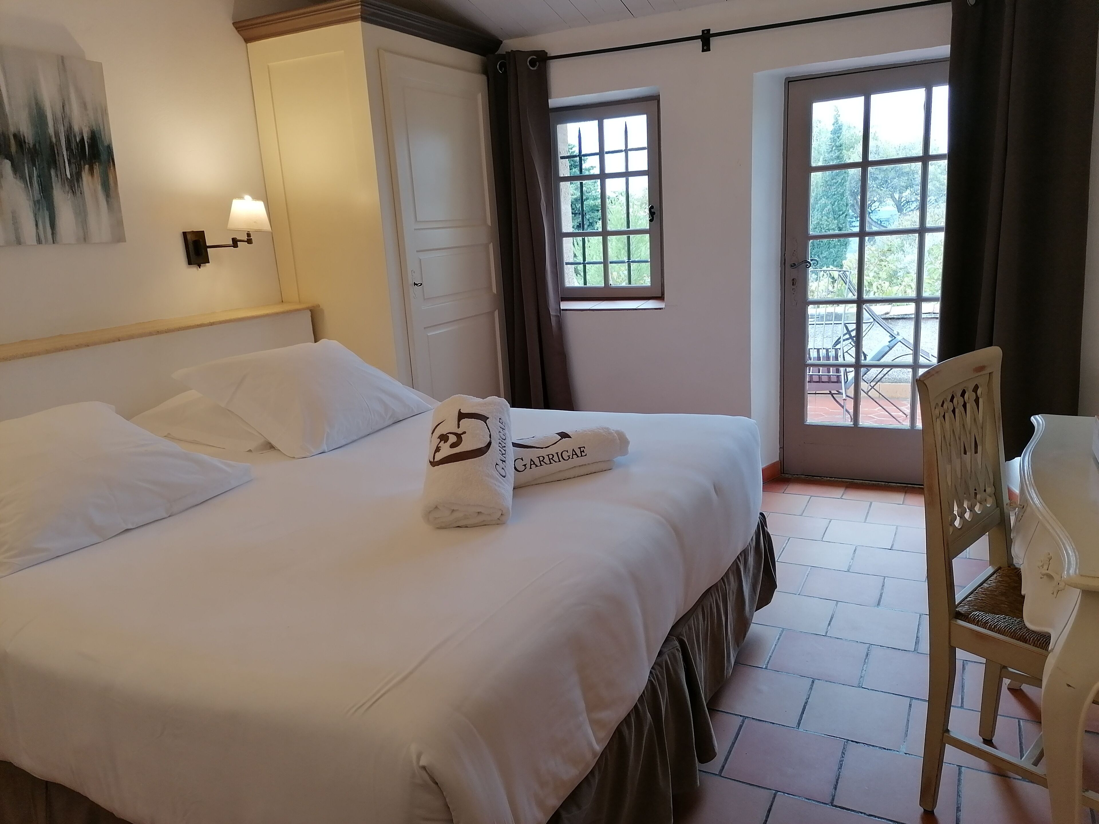 Una cama bien hecha con sábanas blancas y una toalla con un logotipo, una silla de madera y una ventana con cortinas.