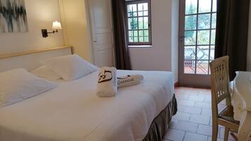 Una cama bien hecha con sábanas blancas y una toalla con un logotipo, una silla de madera y una ventana con cortinas.