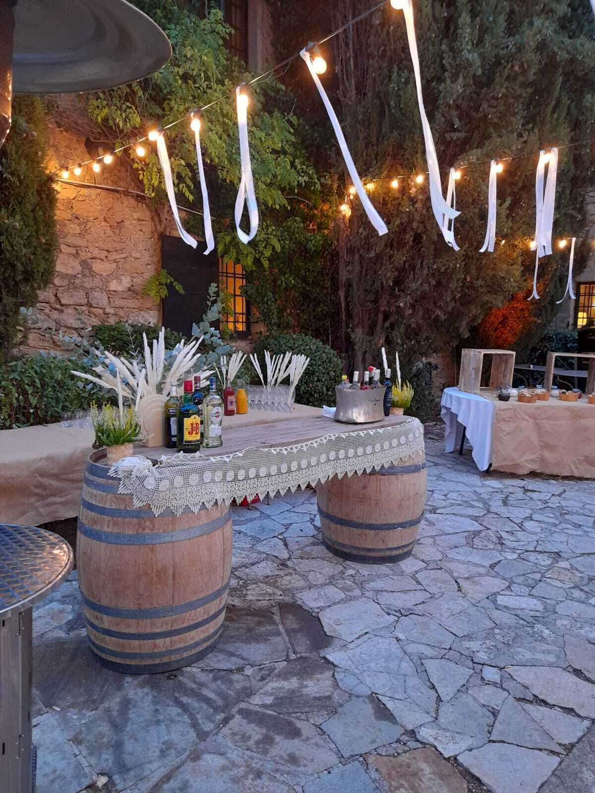 Un patio al aire libre con suelo de piedra, barriles de madera rústica, una mesa cubierta con un mantel de encaje y luces colgantes.