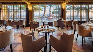 Un restaurant amb taules i cadires rodones, grans finestres amb vista al mar i un terra d'rajola.