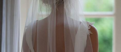 A person in a white dress with a sheer veil, standing in front of a window with a view of greenery.