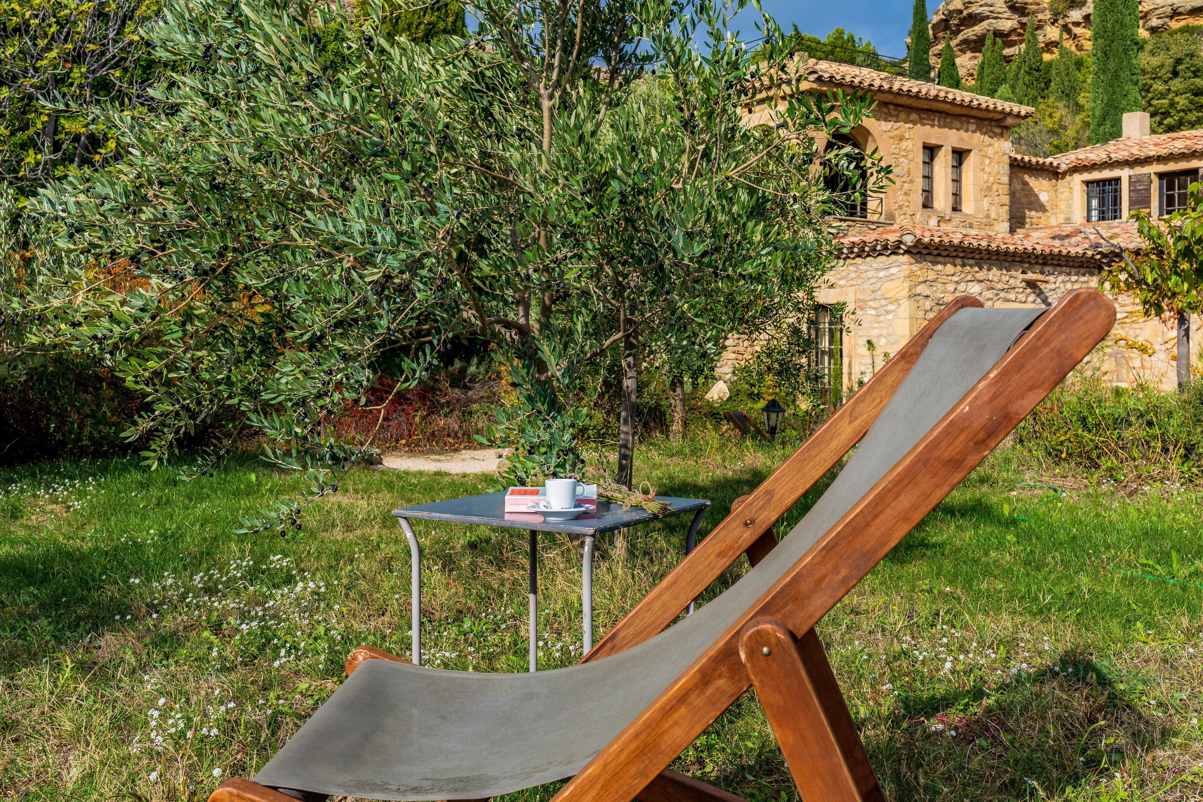 A wooden lounge chair with a metal table and a cup, set in a lush garden with a stone building in the background.