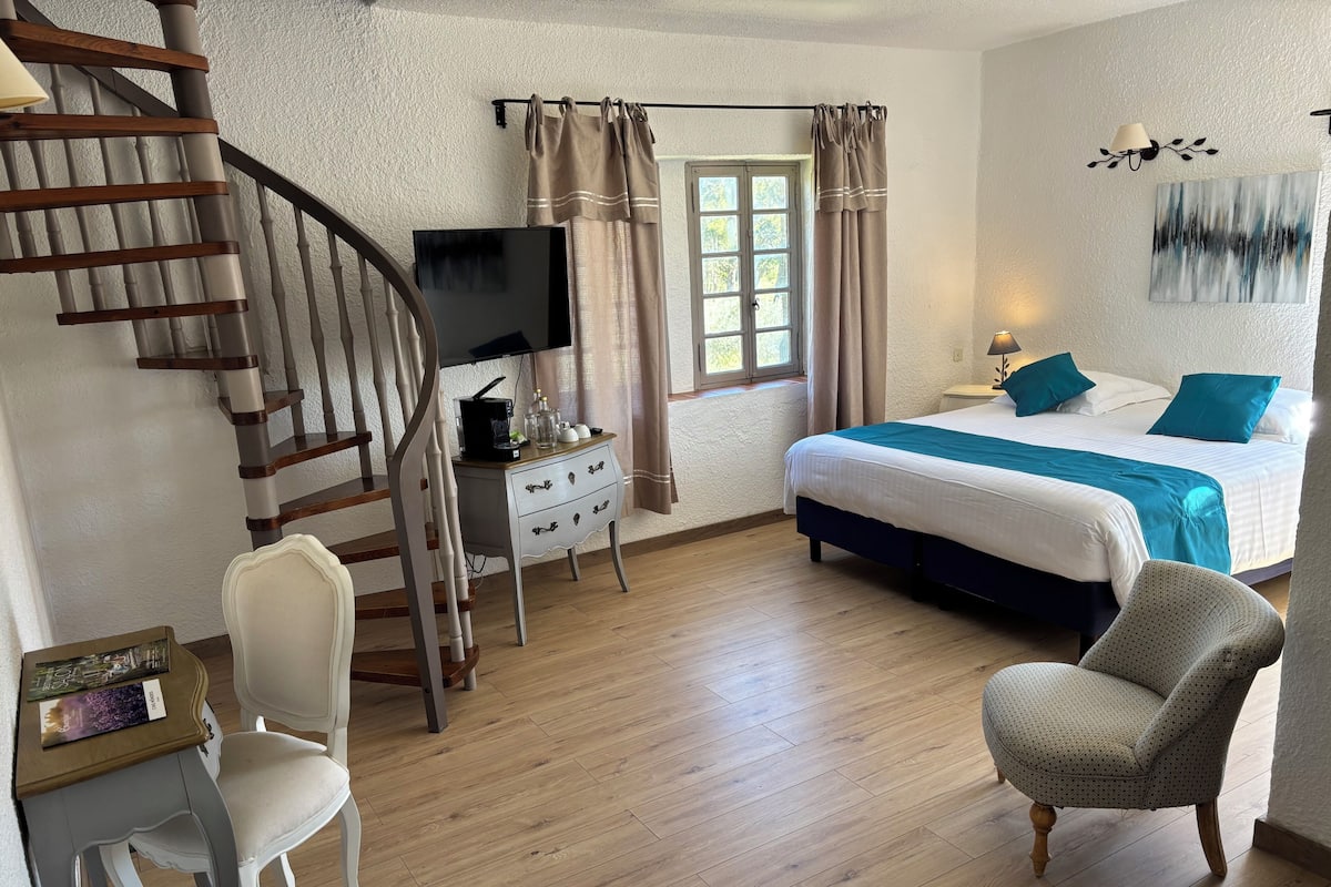 A bedroom with a bed, a desk, a chair, a television, a window with curtains, a staircase, and a wall art piece.