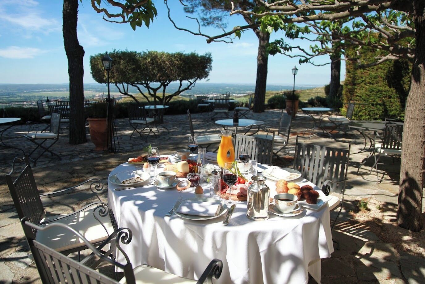 An outdoor dining area with a table set for breakfast, surrounded by chairs and trees, overlooking a scenic view.