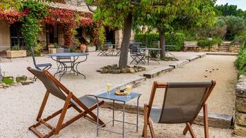 An outdoor seating area with tables and chairs, surrounded by a garden with a stone wall and a tree.