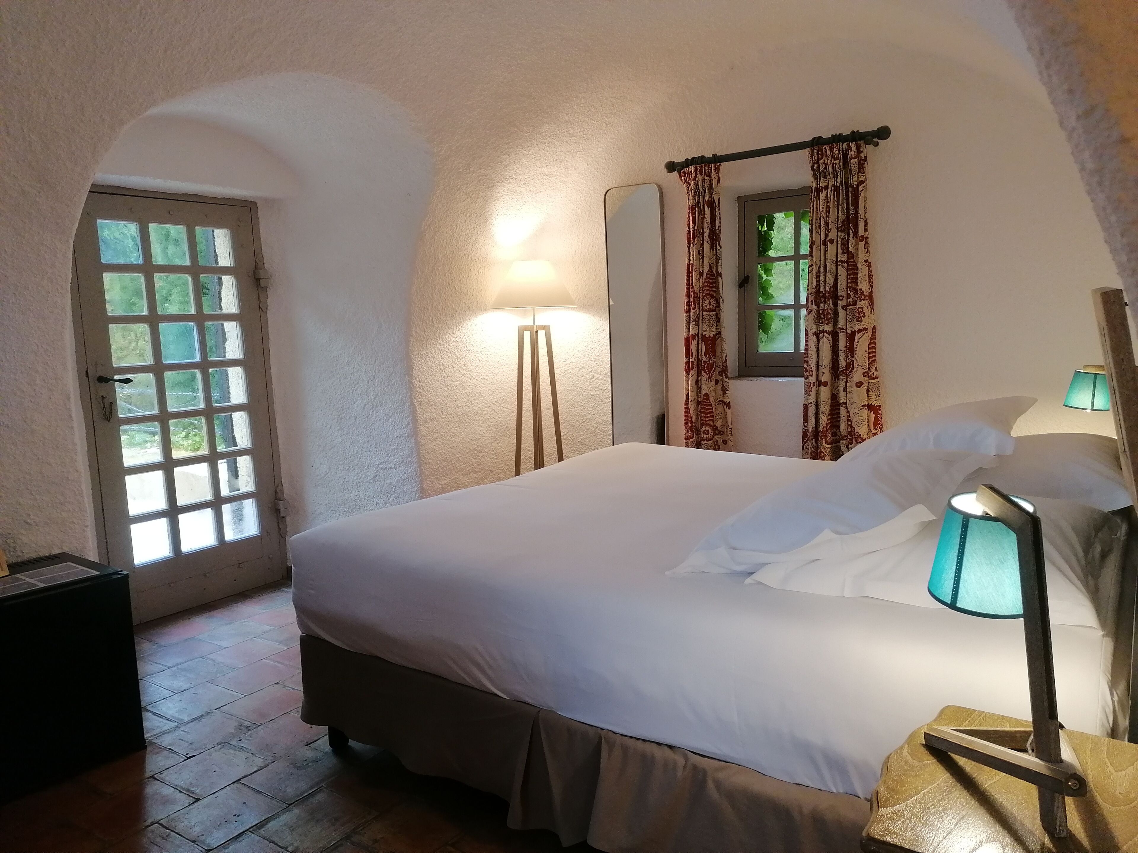 Un dormitorio con una cama grande, una lámpara de noche, un espejo y una ventana con cortinas.