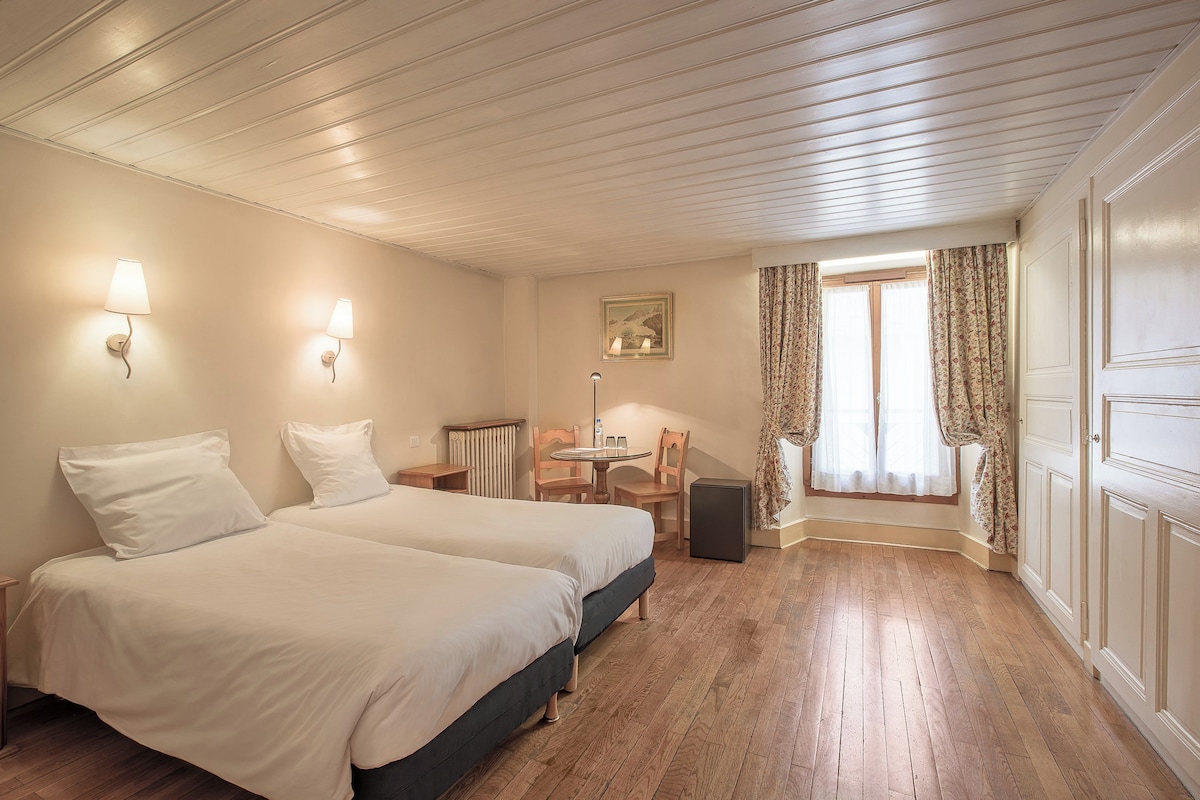 A hotel room with two beds, a small table, chairs, a radiator, and a window with curtains.