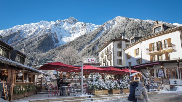 Una montaña cubierta de nieve con cielo despejado, un edificio con el letrero 'L'M', mesas al aire libre con sombrillas rojas y una persona con una mochila.