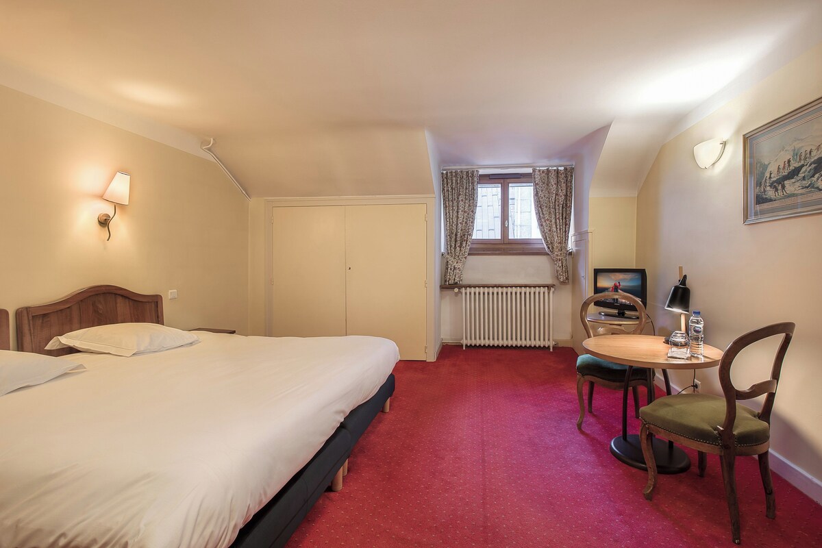 A hotel room with a bed, a desk with a chair, a radiator, and a window with curtains.