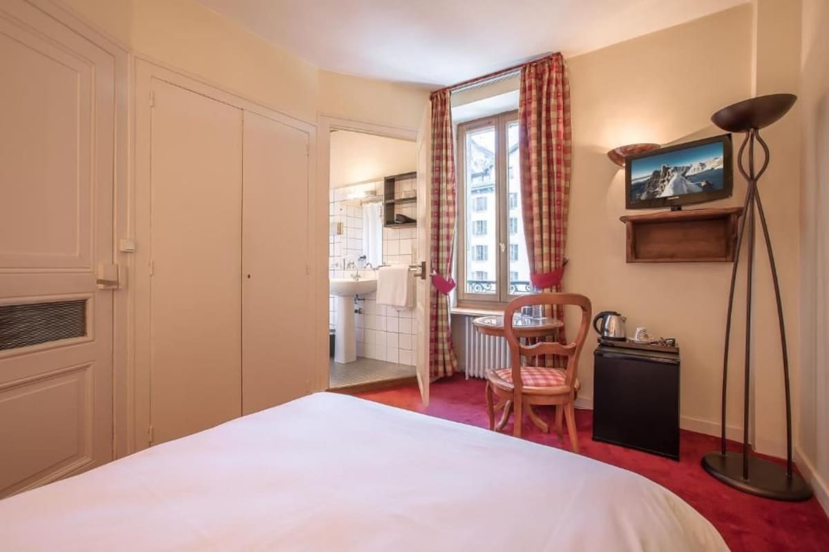 Habitación de hotel con cama, una silla, una mesita, radiador, televisor sobre un mueble y ventana con cortinas.