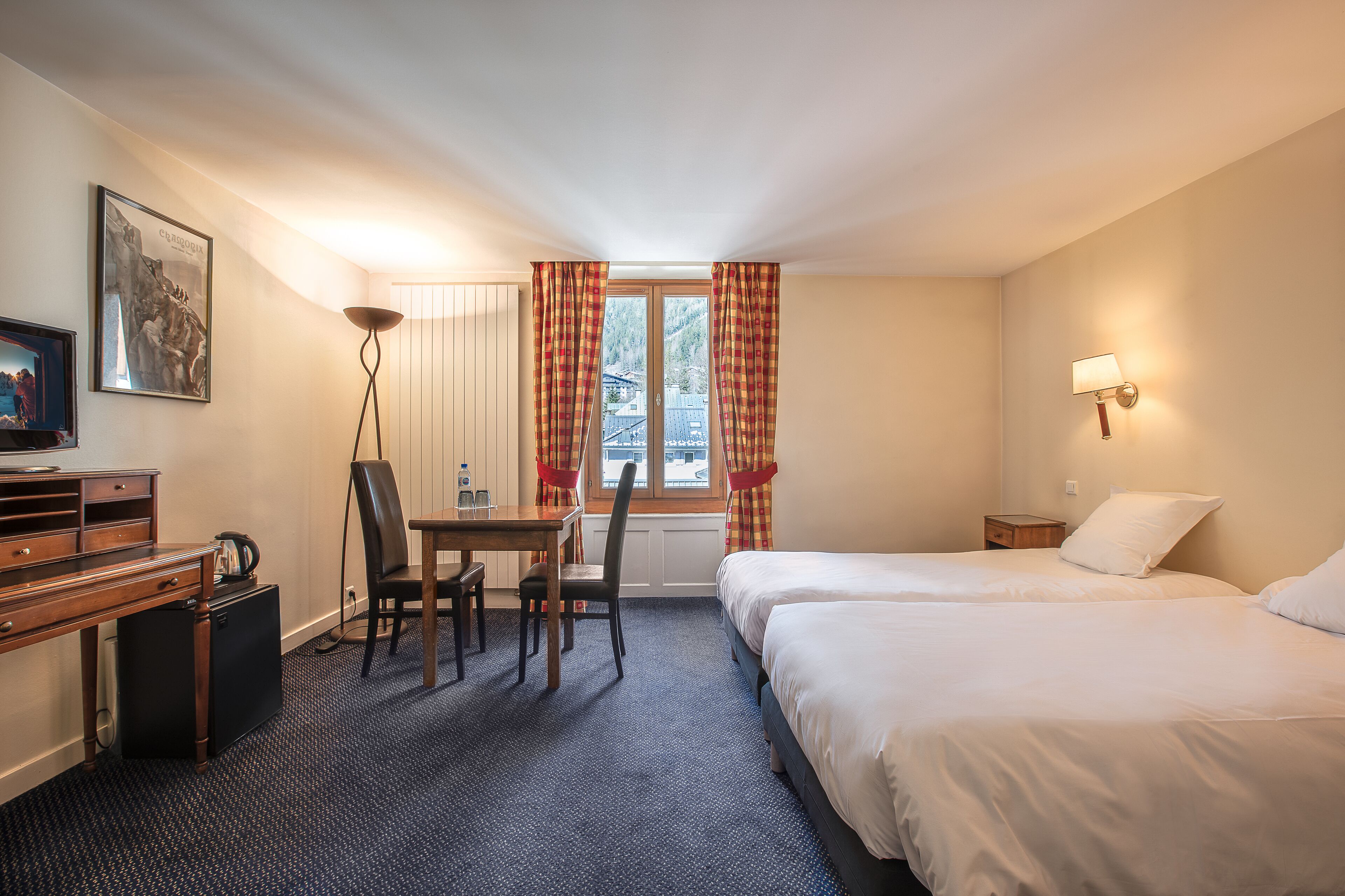Hotelkamer met twee bedden, een bureau met stoel, een kleine tafel, een televisie en een raam met gordijnen.